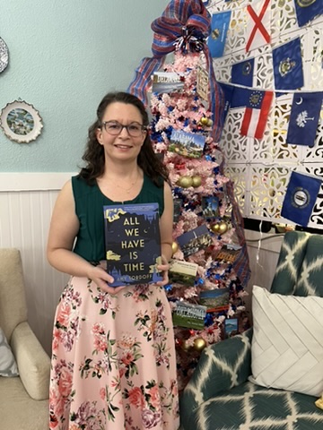 A photo of the author holding her book, All We Have is Time, in front of a pink christmas tree decorated with postcards and glass balls